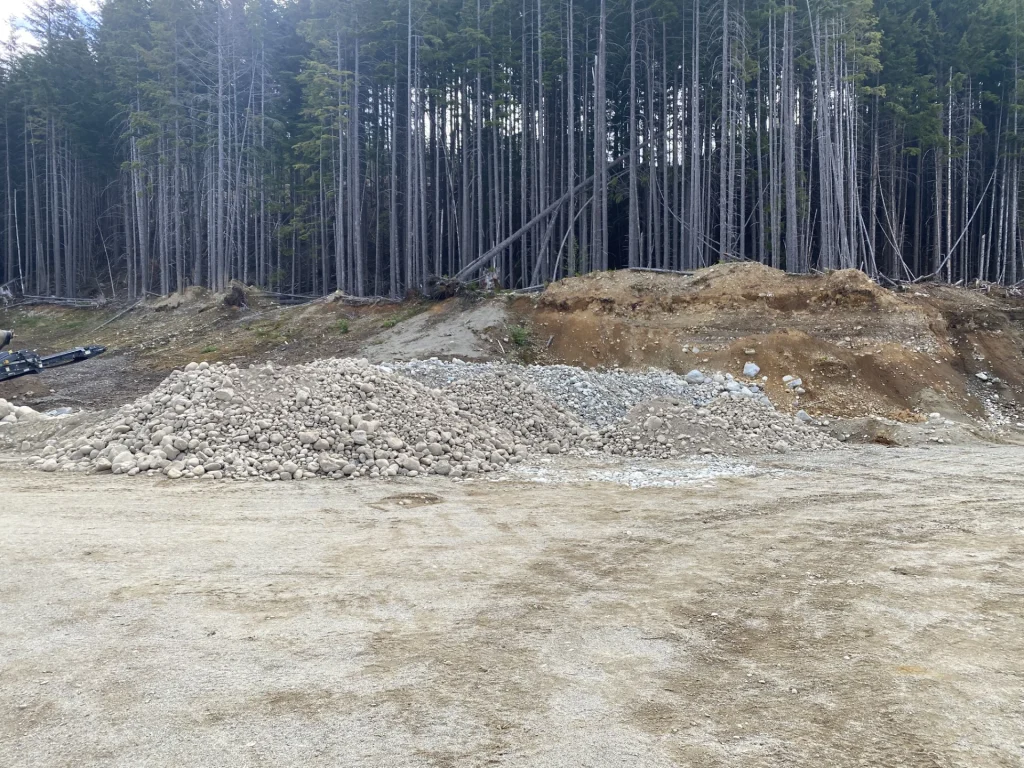 Pile of rounded rock and gravel at an aggregate pit, with exposed earth bank and dense trees behind.