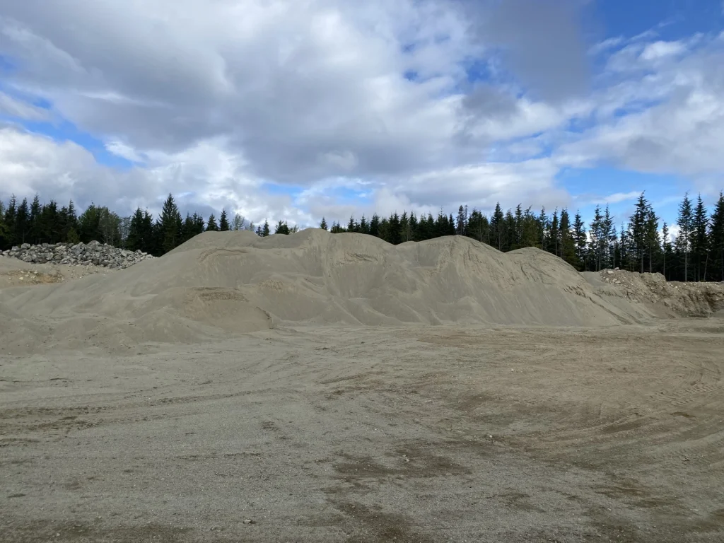 Large stockpile of screened aggregate at an outdoor gravel pit, with forest and cloudy sky in the background.